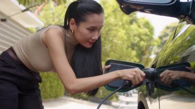 Asya kökenli genç bir kadının portresi otomobil portuna EV ekliyor ve dışarıda kameraya gülümsüyor.