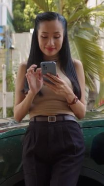 Elektrikli arabanın yanında duran Asyalı kadının dışarıda şarj olurken akıllı telefon kullandığı dikey çekim.