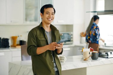 Kameraya gülümseyen Asyalı adamın portresi akıllı telefonuyla mesaj atarken mutfakta duruyordu.