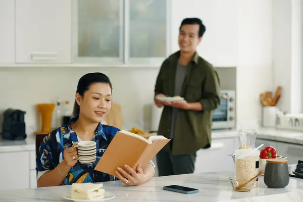 stock image Asian young woman reading a book and drinking coffee while her husband cooking in background