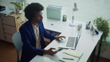 Ofisteki kablosuz bilgisayarın önünde otururken akıllı telefonunu kontrol eden Afrikalı Amerikalı iş adamının yüksek açılı görüntüsü