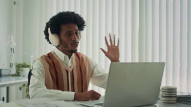 Kulaklıklı siyahi erkek girişimcinin ofis masasında dizüstü bilgisayarın önünde otururken iş arkadaşlarıyla konuşurken orta boy görüntüsü.
