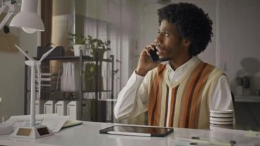 Meşgul Afrikalı Amerikalı erkek yöneticinin cep telefonuyla projenin detaylarını tartışırken ve ofis masasında dijital tablet kullanırken orta boy görüntüsü.