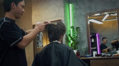 Orta boy Asyalı erkek berberin kuaförde beyaz erkek müşterinin saçını spreyle tararken.