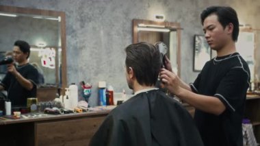 Berber dükkanında çalışırken saç kurutma makineli Kafkas erkek müşterisinin Asya kökenli genç berberi.