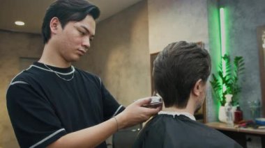 Orta boy Asyalı berber, moda berber dükkanında kırpıcı ile erkek müşterinin saçını buduyor.