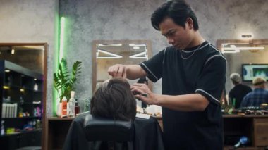 Asyalı berberin, modaya uygun bir salondaki koltukta dinlenen erkek müşterisinin sakalını tıraş edişinin yan görüntüsü