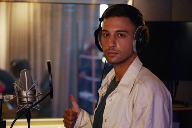 Portrait of vocalist showing thumb up while recording his song in studio clipart