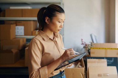 Asyalı bir kadın depoda çalışırken tablet bilgisayarı üzerinde çalışıyor ve çevrimiçi sevkiyatı kontrol ediyor.