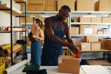 Kadın arka planda mal listesi hazırlarken, erkek kutuları paketlemek için bant kullanıyor