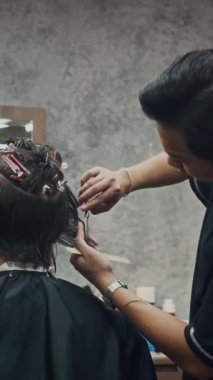 Asyalı berberin erkek müşterinin saçını makasla keserken ve son moda berber dükkanında fırçalarken dikey görüntüsü.