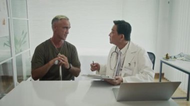 Orta yaşlı Kafkas askeri, Asyalı erkek doktorun klinikteki rehabilitasyon terapisi hakkında konuşmasını dinliyor.