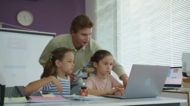Beyaz erkek öğretmenin, küçük kızların okulda ders sırasında dizüstü bilgisayarlarında program yapmalarına yardım ederken çekilmiş statik görüntüleri.