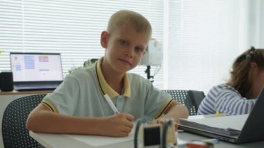 Kafkas kökenli neşeli çocuğun orta boy portresi bilgisayar bilimleri sınıfında test yapıyor ve kameraya gülümsüyor.