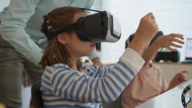 Beyaz erkek öğretmenin okulda bilgisayar dersi sırasında çocuklarla VR gözlüğü kullanırken çekilmiş statik görüntüleri.