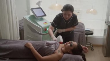 Genç Asyalı güzellik uzmanı klinikteki kadın müşterinin koltuk altı kıllarını temizlemek için lazer ekipmanı kullanıyor.