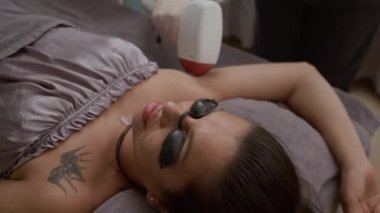 Gözlüklü genç beyaz kadın kuaförde ameliyat sırasında koltuk altı kıllarını lazerle aldırıyor.