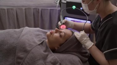 Klinikte tekerlekli ve kırmızı ışık ekipmanıyla maske takan kadın müşterinin orta boy fotoğrafı.