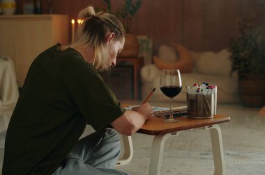 Genç tasarımcının iş yerinde kırmızı şarap kadehiyle oturması ve çizimler yapması.