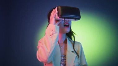 Neon arka planda izole edilmiş VR kulaklıkla video oyunu oynayan zarif kıyafetli genç bir kadının orta boy fotoğrafı.