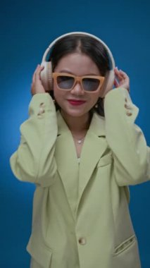 Dikey çekimde güneş gözlüklü genç Asyalı kız müzik eşliğinde neon mavisi arka planda izole bir şekilde eğleniyor.