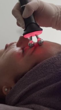 İşlem sırasında genç bir kadının yüzüne kırmızı ışık masaj aleti kullanan bir güzellik uzmanının dikey görüntüsü.