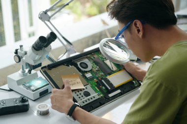 Profesyonel tamirci atölyede bozuk dizüstü bilgisayarı tamir ederken büyüteç kullanıyor