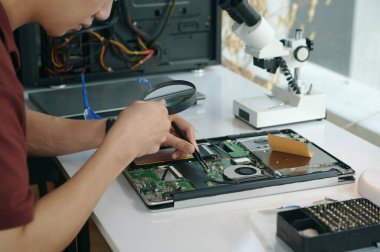 İşyerinde otururken bilgisayarın ana kartını büyüteçle inceleyen teknisyen.