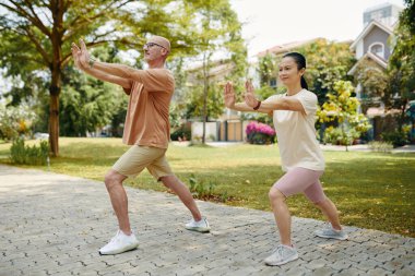 Çevrelerinde ağaçlar ve yeşillikler olan açık hava parkında Tai Chi pratiği yapan iki kişi, dikkatle hareket ettiklerini gösteriyorlar, yaşlı adam ve Asyalı kadın da dahil.