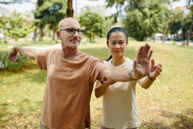 Huzurlu parkta Tai Chi pratiği yapan yaşlı bir adam ve Asyalı bir kadın. Etrafı yeşilliklerle çevrili. Rahat, sakin ifadelerle, günlük kıyafetleriyle, kesin hareketlere odaklanmış.