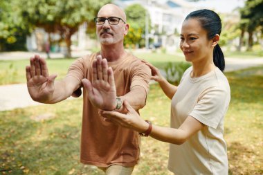 Tai Chi 'yi eğitmen duruşu ile parkta pratik yapan kadın ve erkek. Açık hava aktivitesi denge, rahatlama ve refah odaklı, arka planda ağaçlar ve yeşillik ile