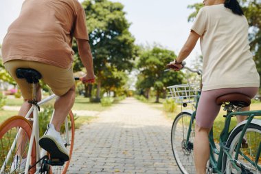 Güneşli bir günde, verimli ağaçlarla çevrili asfalt yol boyunca giden iki bisikletçi. Her iki katılımcı da günlük kıyafetlerini giyip dışarıda eğlenirken sohbet ediyorlar.