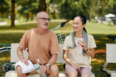 Sırıtkan Kafkas adam ve Asyalı kadın parkta bankta oturuyor, arka planda bisikletler ve içki şişeleriyle su içerken sohbetin keyfini çıkarıyorlar.