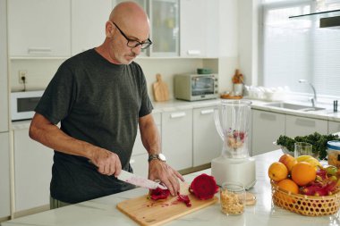 Mutfakta kıdemli bir adam ahşap tahtada taze meyve keserken sağlıklı gıda hazırlama rutinini gösteren sağlıklı malzemelerle çevrili bir blender kullanıyor.