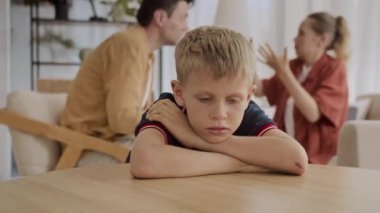 Medium shot of frustrated Caucasian teenage kid sitting at table while his parents having quarrel in background at home