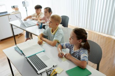 Dört genç öğrenci, arka planda büyük pencereleri olan modern masalarda otururken dizüstü bilgisayarlar ve defterler kullanarak sınıf etkinliğine katılıyorlar.