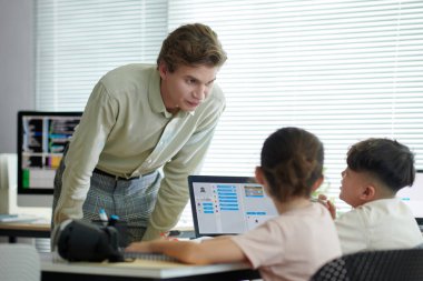 Öğretmen bilgisayarların önünde eğitici materyalleri açıklarken iki öğrenciyle takılıyor. Son teknolojiyle donatılmış sınıf odası, etkileşimli bir ortam yaratıyor.