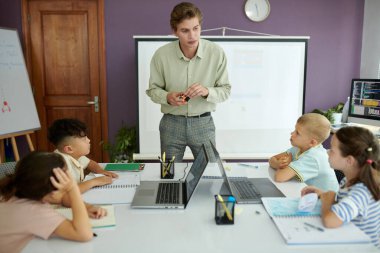 Modern sınıf ortamında ders sırasında dört çocuğa rehberlik eden öğretmen, onları laptoplarla ve büyük masadaki defterlerle elle etkinlikle meşgul ediyor.