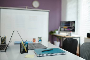 Boş ofis masasında iki dizüstü bilgisayar, not defteri, kalem ve iyi ışıklandırılmış mor duvar ve pencere perdeleri olan kırtasiye konteynırı var. Arkaplan unsurları arasında ofis ekipmanları ve ev bitkileri