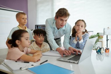 Sınıfta kitaplarla ve kağıtlarla çevrili, camdan gelen parlak ışıkla çocuklara rehberlik eden öğretmen. Çocuklar dikkatle ekranı izliyor