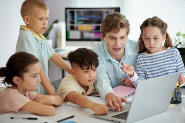 Bir grup çocuk ve öğretmenleri, arka planda eğitim ekranlarıyla birlikte sınıf ortamında dizüstü bilgisayarın etrafında ders üzerinde işbirliği yapıyorlar.