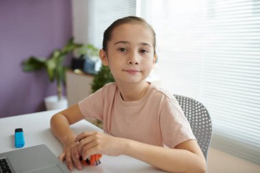 Portrait of young girl sitting by desk with laptop and toy car, looking straight at camera, having short brown hair pulled back, natural light illuminating room with plants in background clipart