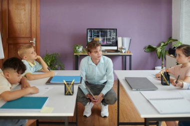 Sınıfta grup tartışması sırasında genç öğrencilerle ilgilenen öğretmenler ve çocuklar eğitim araçları ve malzemelerle masalarda oturuyorlar.