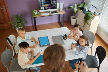 Çocuklar sınıf masasında oturuyor, çeşitli aktivitelere katılıyorlar. Yeşil bitkiler ve kırtasiye malzemeleri odayı dekore ederek, canlı bir öğretim ortamı yaratıyor.
