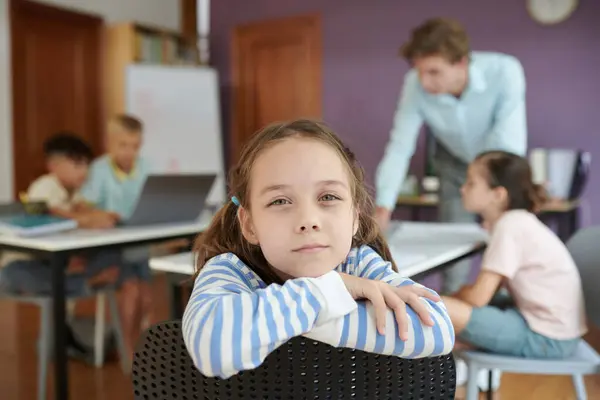 stock image Portrait of young girl sitting in classroom setting with other students and teacher in background, showing focused expression Study materials and laptops on desks