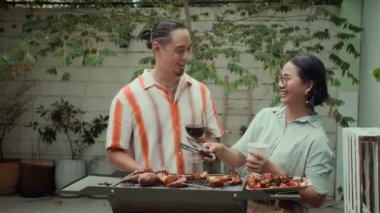 Orta halli, etnik çeşitlilikte çiftin ızgara yaptığı, kırmızı şarap içtiği ve dışarıda konuştuğu bir fotoğraf.