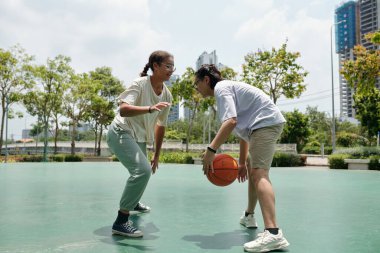 Güneşli bir günde çevresi yeşillik ve şehir binalarıyla çevrili açık sahada basketbol oynayan iki genç.