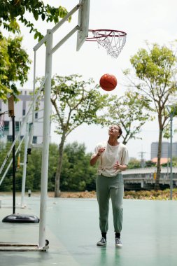 Dışarıda basketbol oynayan, güneşli bir günde çemberden düşen ve arka planda ağaçlar ve binalar olan topu yakalamaya hazırlanan bir adam.