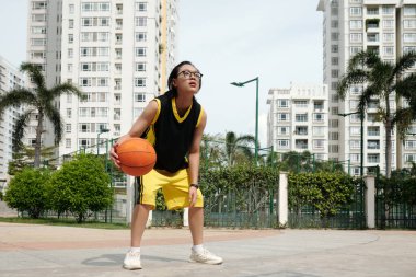 Basketbol gözlüğü takan ve şehir sahasında dikilen genç, açık gökyüzü altında konut binaları ve yeşilliklerle çevrili.