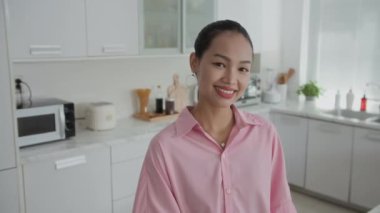 Ev mutfağındaki kameraya gülümseyen Asyalı kadın kıyafetinin orta boy portresi.